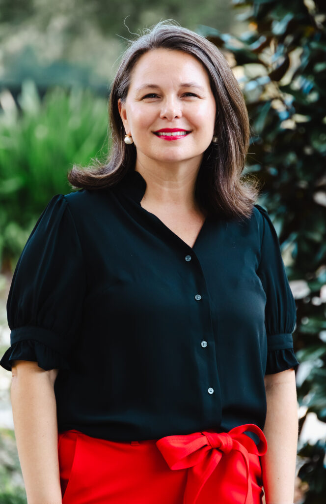 Arica Heise portrait. Woman in black blouse and red pants stands outdoors, smiling.