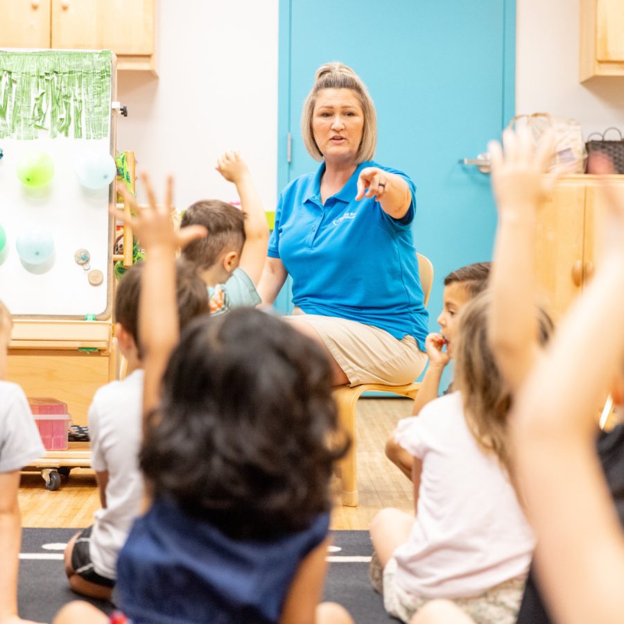 A preschool teacher calls on her students as many raise their hands.