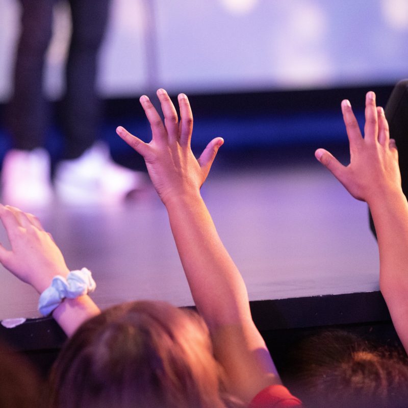 Students raise their hands in worship during an event in the auditorium at The Rock School.