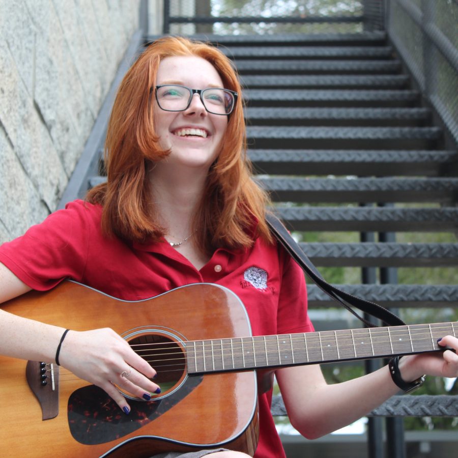 A High School Student with red hair and glasses playing a guitar