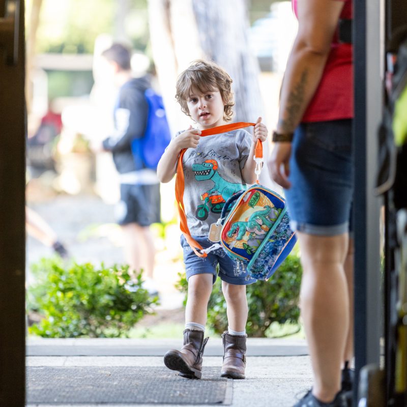 Preschool student walking into school holding a backpack.