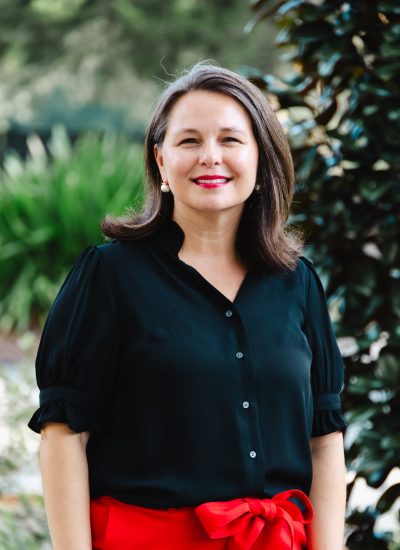 Arica Heise, smiling woman in a black blouse and red pants, outdoor portrait.