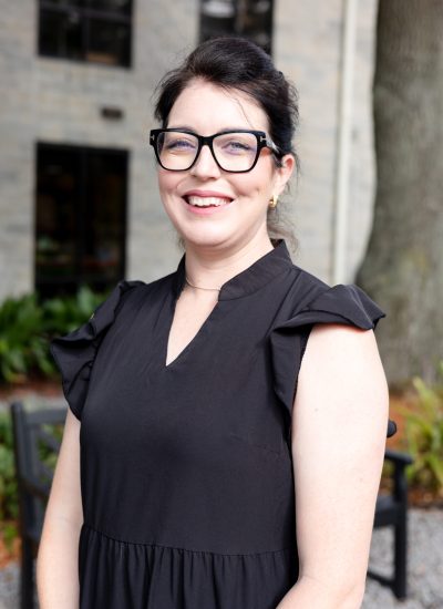 Jolyn Caddell smiles wearing glasses and a black dress. Headshot outdoors.