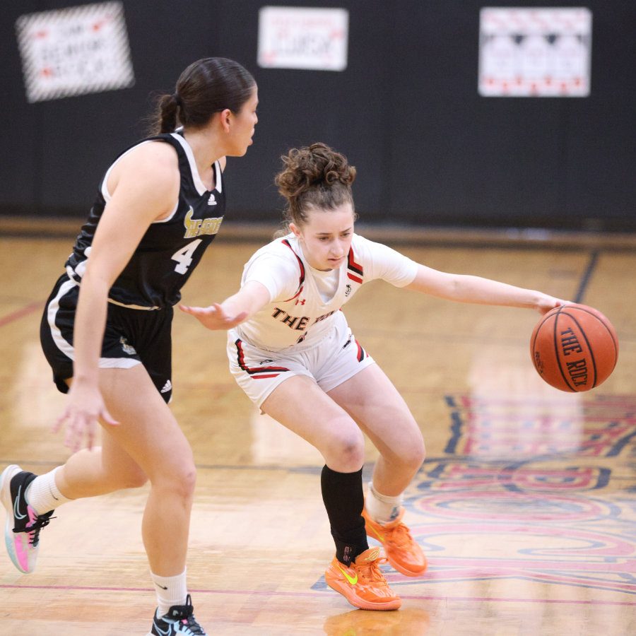 Two female high school athletes compete in a game of basketball whilst one is dribbling towards the basketball hoop.